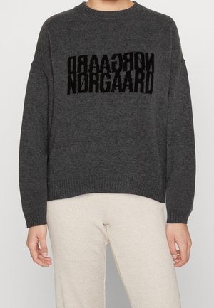 Personne portant un pull à manches longues gris foncé avec un texte noir miroir "NORGAARD" sur la poitrine et un pantalon beige clair côtelé.
