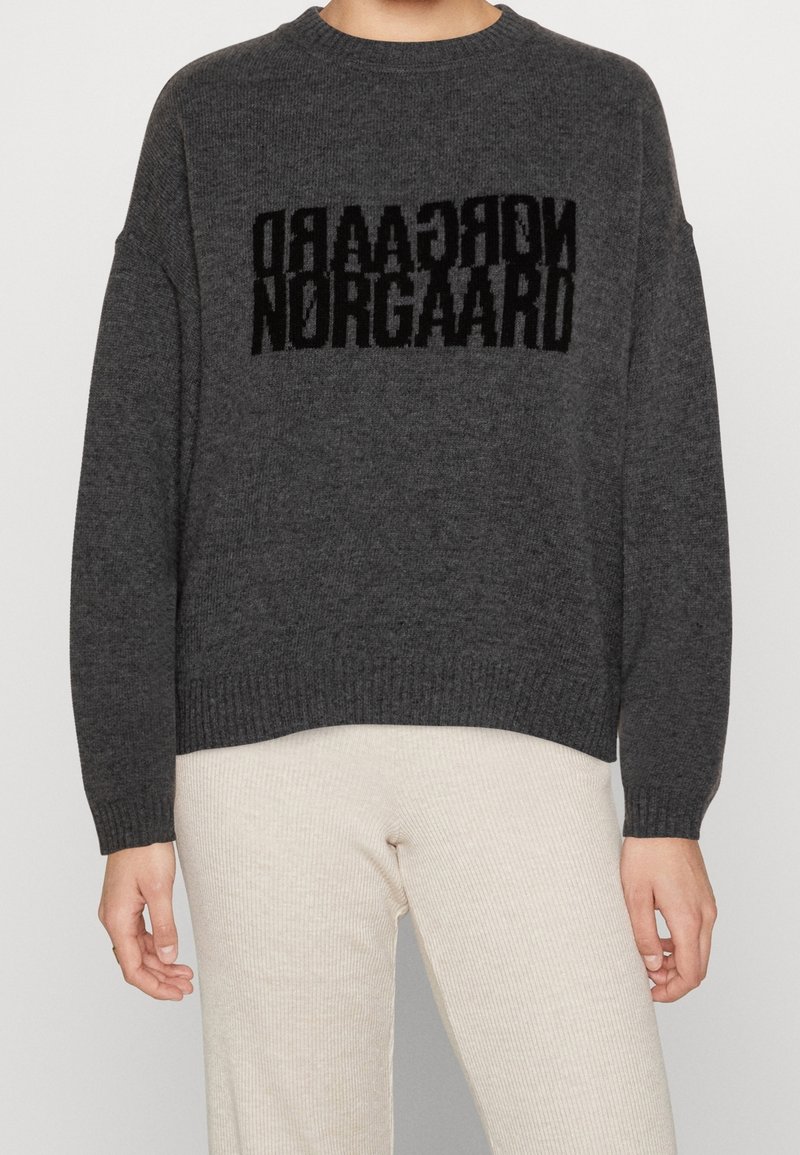 Personne portant un pull à manches longues gris foncé avec un texte noir miroir "NORGAARD" sur la poitrine et un pantalon beige clair côtelé.