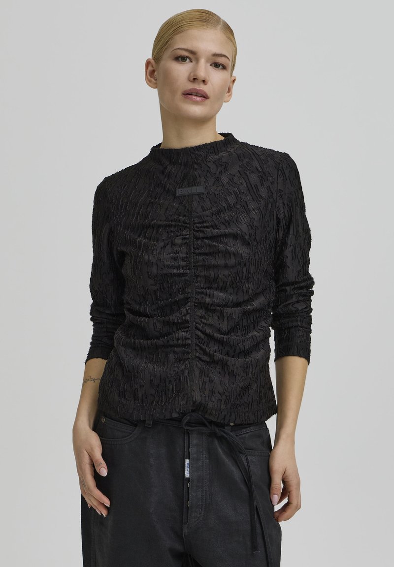 Fekete, hosszú ujjú felső texturált anyagból, gyűrt mintával és finom logóval az elülső részen. Sima, bordázott nyakkivágással.