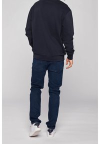 Man i mörkblå jeans, marinblå sweatshirt och vita sneakers står med ryggen mot kameran på en enkel grå bakgrund.