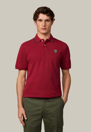 Camisa polo roja de algodón, con un cierre de dos botones, rayas de contraste en el cuello y un parche del logo en el lado izquierdo del pecho.