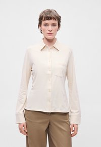 Ανοιχ�τό μπεζ πουκάμισο με μακριά μανίκια, κούμπωμα με κουμπιά στη μπροστινή πλευρά, κολάρο τύπου πόντ και μία τσέπη στο στήθος. Λείος υφή και ελαφριά γυαλάδα.