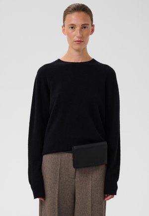 Donna con i capelli pettinati all'indietro che indossa un maglione nero e pantaloni marroni, che tiene una pochette nera in vita davanti a uno sfondo semplice.
