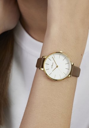 Runde goldene Uhr mit einem weißen Zifferblatt, dünnen goldenen Stundenmarkierungen und einem braunen Lederarmband, die auf einem hellen Arm liegt. Einfaches und elegantes Design.