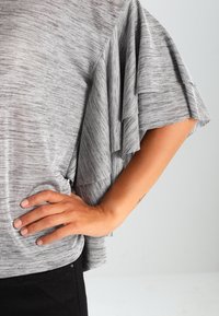 Grijze, gestructureerde top met gelaagde rucheslitzakken. Close-up van het kledingstuk met een zwarte tailleband en een hand op de heup.
