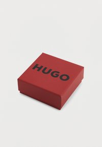 Scatola regalo quadrata in cartone rosso con logo nero "HUGO" sulla superficie superiore, forma quadrata con una texture liscia e senza motivi visibili.
