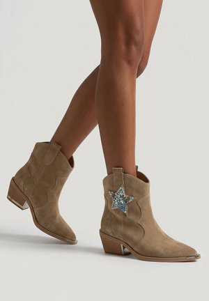Beine, die hellbraune Wildleder-Stiefeletten mit niedrigen Holzabsätzen und einem dekorativen blauen Paillettenstern an der Außenseite tragen.