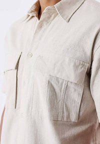 Camisa de lino beige de manga corta, con dos bolsillos en el pecho, cierre de botones y un acento bordado cerca del cuello. Tela texturizada.