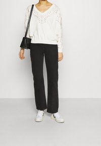 Witte lange mouw sweater met V-hals en decoratieve oogpatronen, gecombineerd met zwarte straight-leg jeans en witte sneakers. Zwarte crossbody tas.