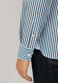 Striped button-up shirt with green, blue, and white vertical lines, featuring a cuff with two pearl buttons and a rounded hem.