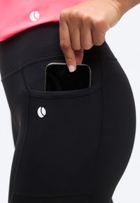 Χέρι που βάζει smartphone στην πλαϊνή τσέπη μαύρου κολαν, φορώντας ροζ μπλούζα, με λευκό λογότυπο στην τσέπη και στο πουκάμισο.