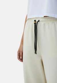 Lichtbeige joggers met een zachte textuur, voorzien van een elastische tailleband en zwarte trekkoorden met gouden uiteinden.