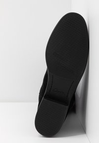 Semelle de chaussure noire avec une prise texturée et un talon bas, inclinée contre un fond blanc en coin, montrant le nom de la marque embossé sur la semelle.