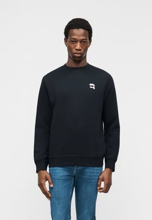 Homme portant un sweat-shirt noir avec un petit motif blanc sur la poitrine et un jean bleu, debout devant un fond clair uni.