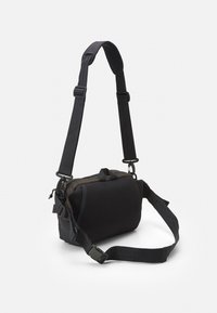 Černá crossbody taška se strukturovaným síťovým předním dílem, nastavitelným popruhem a zipovým uzávěrem. Má kompaktní, obdélníkový tvar.