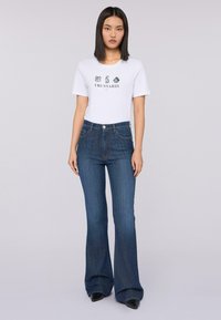 Donna in jeans a zampa scuri, scarpe nere a punta e t-shirt bianca a maniche corte con il testo e il logo "TRUSSARDI" davanti.