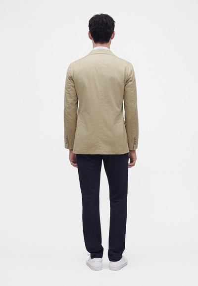 Man staat met de rug naar ons toe, draagt een beige blazer, donkere broek en witte schoenen tegen een effen witte achtergrond.