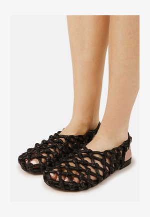 Pieds portant des sandales plates noires à motif noué tissé, avec orteils ouverts et une bride rose au talon, sur un fond uni.