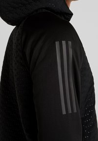 Chaqueta negra con tejido texturizado, diseño acolchado y capucha. Presenta tres franjas paralelas en el hombro con un acabado mate.