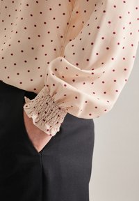 Blusa a maniche lunghe di un delicato rosa chiaro con pois bordeaux. Presenta polsini elasticizzati arricciati e una texture liscia e morbida. Indossata con pantaloni neri.