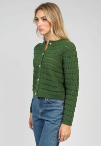 Cardigan en tricot vert avec un motif horizontal, col rond et boutons en or. Design ajusté, associé à un jean en denim bleu.