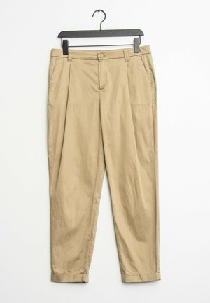 Pantaloni di cotone beige con pieghe, passanti per cintura, tasche laterali e orli risvoltati appesi a una gruccia di plastica nera.