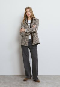 Blazer de pata de gallo marrón y beige con cuello con muesca, sobre una camiseta gráfica gris y pantalones anchos grises, combinados con zapatos marrones.