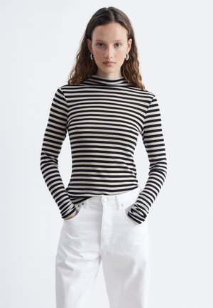 Jeune femme aux longs cheveux bouclés portant un haut à manches longues rayé noir et blanc et un pantalon blanc, les mains dans les poches.