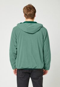 Grüne Fleece-Hoodie mit strukturiertem Stoff, Kordelzugkapuze und langen Ärmeln, über einem dunklen Shirt getragen und mit schwarzen Hosen kombiniert.