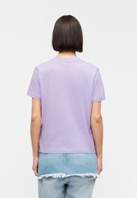 Persoon met kort donker haar, gekleed in een effen lavendelkleurig T-shirt en een lichtblauwe versleten spijkerrok, met de rug naar de kijker tegen een witte achtergrond.