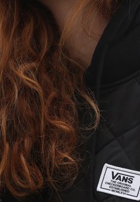 Czarna pikowana kurtka z białą naszywką z logo Vans, kontrastującą teksturą materiału oraz kręconymi, rudo-brązowymi włosami na pierwszym planie.