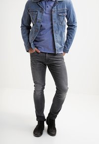 Džínová bunda v světle modré barvě, šedé polo tričko, slim šedé džíny a černé boty. Bunda má dvě náprsní kapsy a klasický límec.