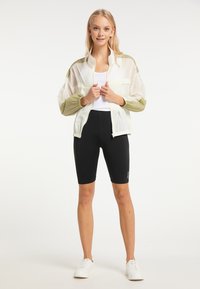Giacca leggera bianca e verde oliva con maniche trasparenti, abbinata a pantaloni ciclismo neri e sneakers bianche.