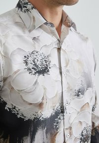 Chemise à manches longues en tissu lisse, avec un motif floral blanc sur fond noir, dotée d'un col classique et de boutons.