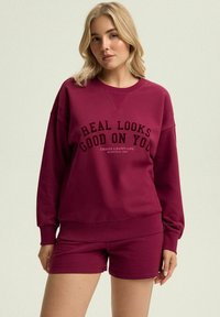 Bordowy zestaw składający się z bluzy i szortów z nadrukowanym czarnym tekstem. Bluza ma okrągły dekolt i ściągacze; miękka tekstura materiału.