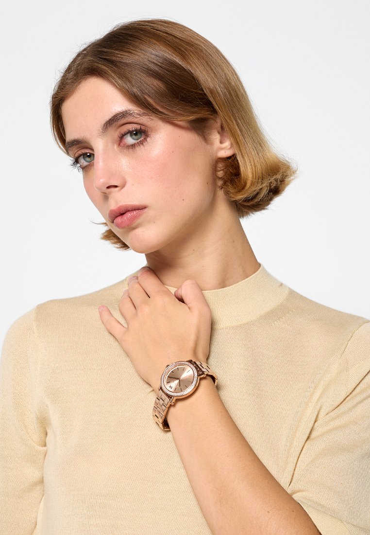 Roségoldene Uhr mit einem metallischen Armband, rundem Ziffernblatt und einem mit Kristallen verzierten Gehäuse. Am Handgelenk eines in hellbeige Pullover gekleideten Arms getragen.