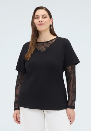 Nainen pukeutunut mustaan yläosaan, jossa on läpikuultavat kukkakirjotut pitsihihat ja kaula-aukko, yhdistettynä valkoisiin housuihin, seisoo yksinkertaisen vaalean taustan edessä.