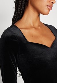 Haut à manches longues en velours noir avec un décolleté carré, présentant une texture lisse et une coupe ajustée, mettant en valeur la zone de la clavicule.