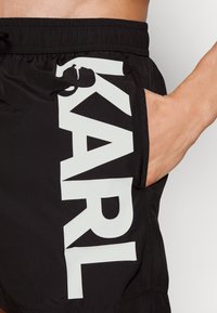 Juoda maudymosi šortai su elastinga juostele, su dideliu baltu "KARL" logotipu šone ir ranka kišenėje. Lygus paviršius.