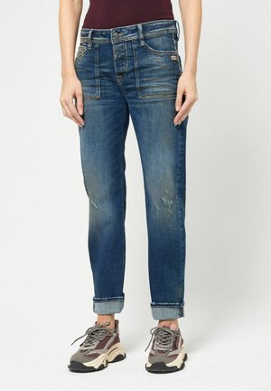Person trägt ausgewaschene blaue Jeans mit aufgekrempeltem Saum und klobige graue und pinke Sneakers und steht vor einem schlichten weißen Hintergrund.