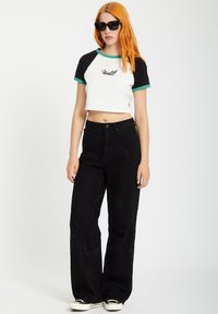 Gekürztes weißes T-Shirt mit schwarzen Ärmeln und grünen Akzenten, ausgestattet mit einem Flügel-Design, kombiniert mit lockeren schwarzen Jeans und schwarzen Sneakers.