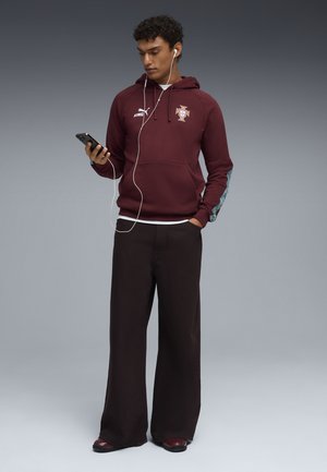 Młody mężczyzna w bordowej sportowej bluzie z kapturem i szerokich czarnych spodniach, patrzący na telefon, noszący słuchawki, stoi na szarym tle.