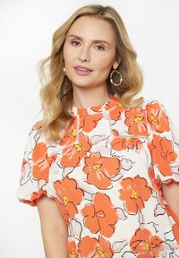 Blumige Bluse mit orangefarbenen und gelben Blumen auf weißem Hintergrund, ausgestattet mit kurzen Puffärmeln und einem gerafften Ausschnitt.