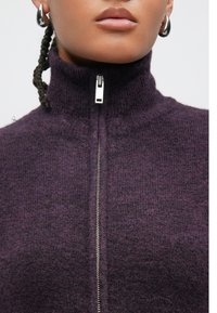 Vijoličen pulover z zadrgo in visokim ovratnikom, s kovinsko zadrgo in teksturiranim pletenim vzorcem. Mehki material s prijetnim videzom.