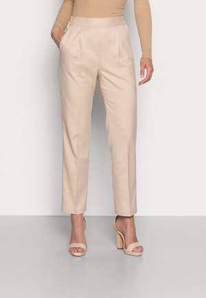 Femme portant un pantalon beige taille haute avec jambes droites et poches latérales, associé à des sandales nude à talons blocs ouverts à l’avant et un top à manches longues côtelé.