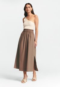 Brun linne maxi kjol med elastisk midja och sido slits, i kombination med en krämfärgad top med en axel och beige flätade slidesandaler.