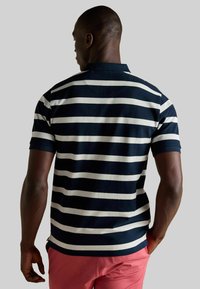 Jūras spēku un baltā svītraina polo krekls ar teksturētu audumu, klasisku apkakli un īsām piedurknēm, kombinēts ar rozā šortiem.