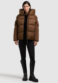 Giacca a vento marrone con cappuccio, caratterizzata da un design trapuntato, zip frontale e accessori neri. Abbinata a leggings neri e stivali al ginocchio.