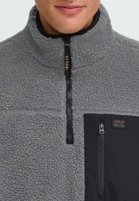 Grauer Fleece-Halbzip-Pullover mit einem kontrastierenden schwarzen Einsatz; ausgestattet mit einer Reißverschlusstasche und Branding; weiche Textur mit erhöhten Fasern.