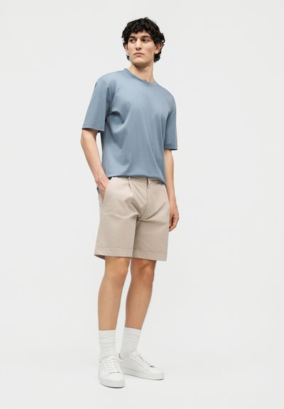 Jeune homme debout, les mains dans les poches, portant une chemise à manches courtes bleu clair, un short beige, des chaussettes blanches et des baskets blanches.